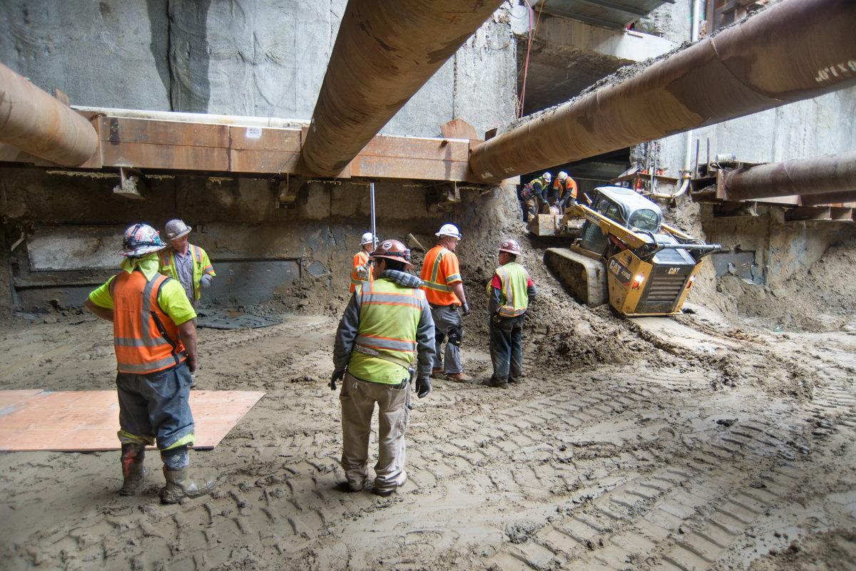 Construction workers in safety gear transfer materials beneath elevated concrete structures at a subway station construction site.