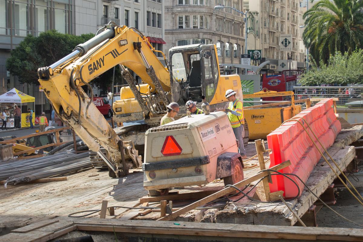 Yellow excavator and construction equipment on wooden deck at Union Square Garage construction site with red traffic barriers and city buildings in background.