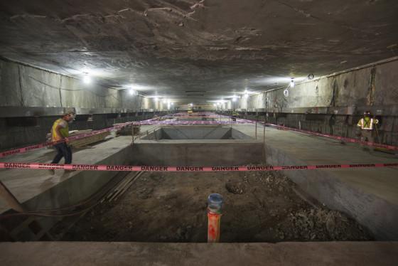 Underground transit station under construction with concrete floor slab, safety barriers, and work lighting.