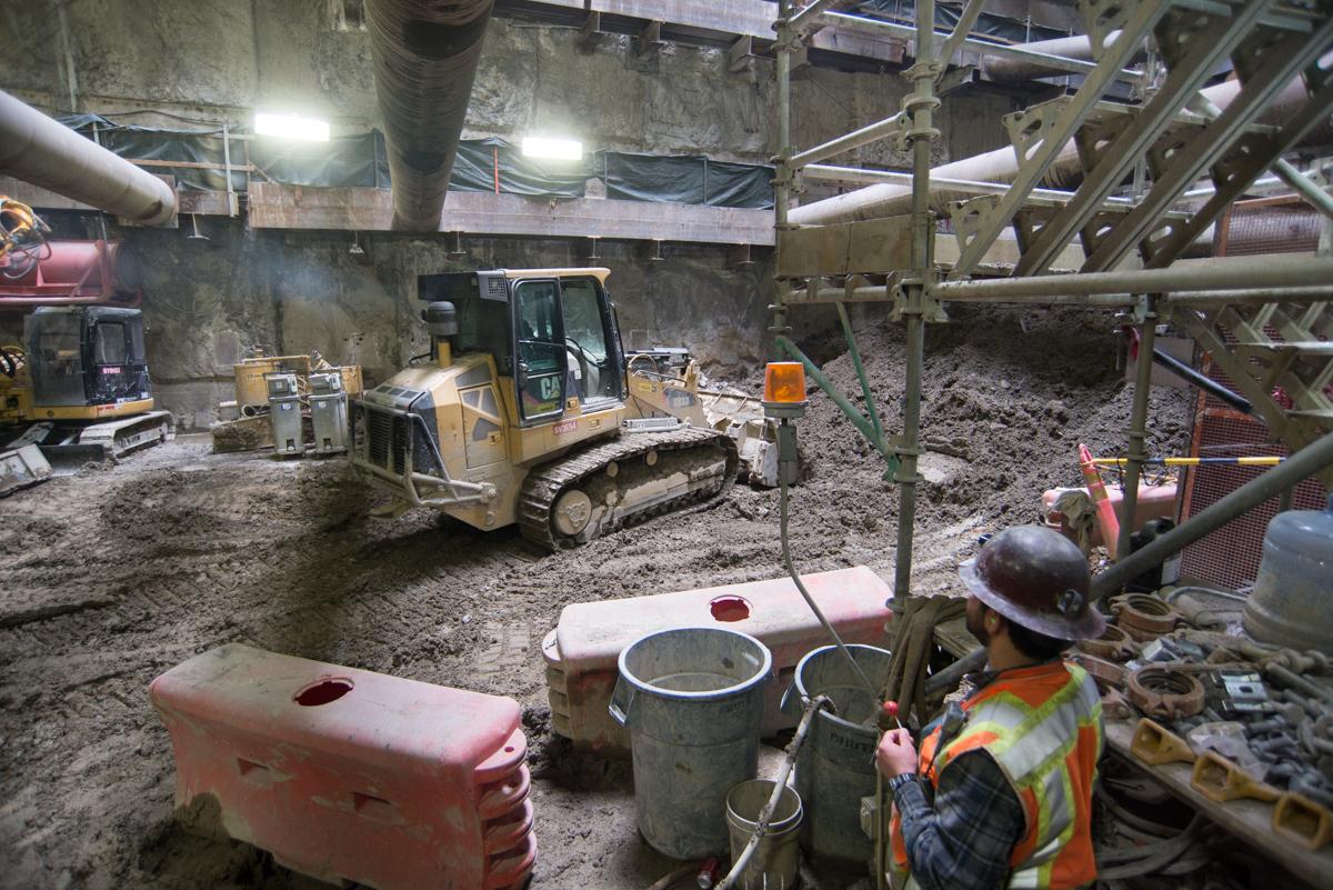 Construction worker in safety gear operates tracked excavator in underground station cavern during platform excavation work.
