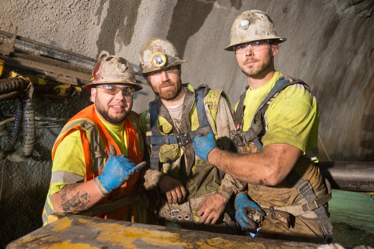 Three construction workers in hard hats and safety vests stand in an underground tunnel next to excavation equipment.