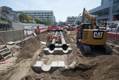 Construction site with excavator, sewer pipes, and heavy equipment in urban setting.
