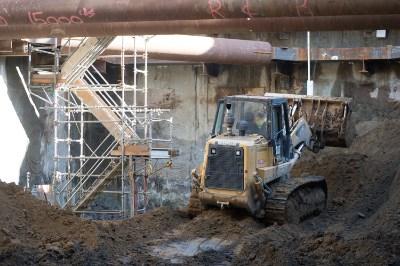 Bulldozer removing soil from station excavation site with temporary steel shoring and industrial structure visible.