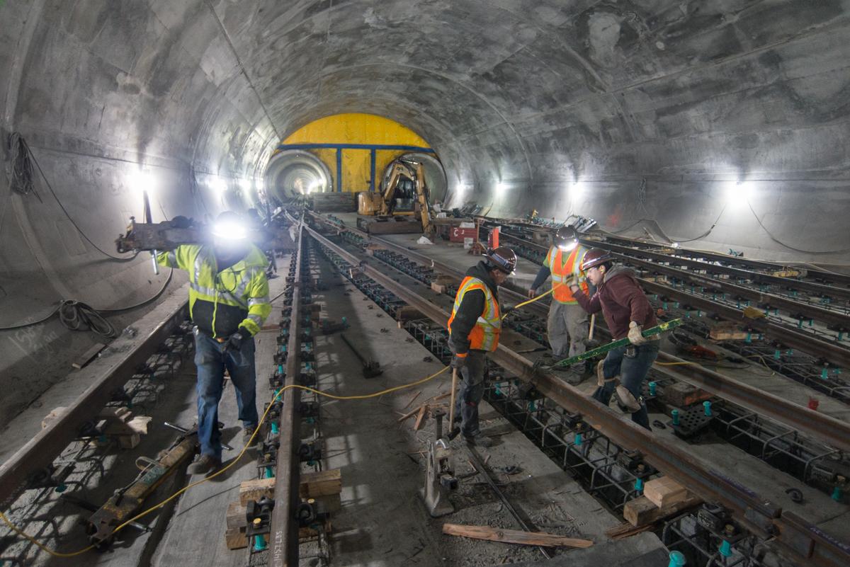 Workers install rail sections in underground tunnel with yellow equipment and LED lighting.