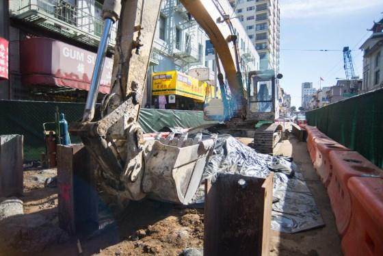 Excavator removing material from underground shaft on Stockton Street construction site surrounded by barriers and urban buildings.