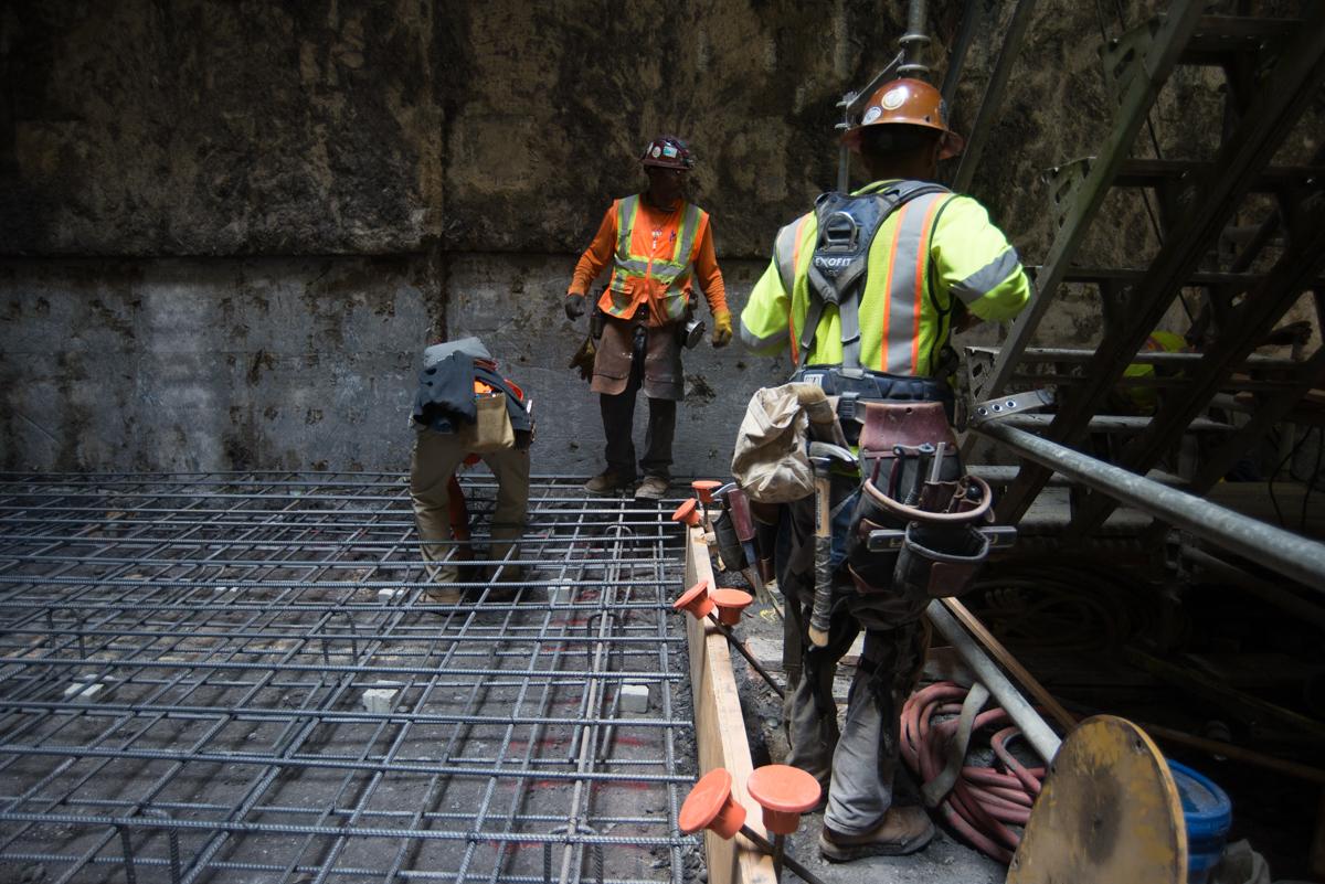 Construction workers install steel rebar reinforcement on the floor of an underground transit station excavation site.