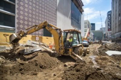 Yellow excavator removing soil and debris at Union Square Garage construction site with downtown buildings in background.