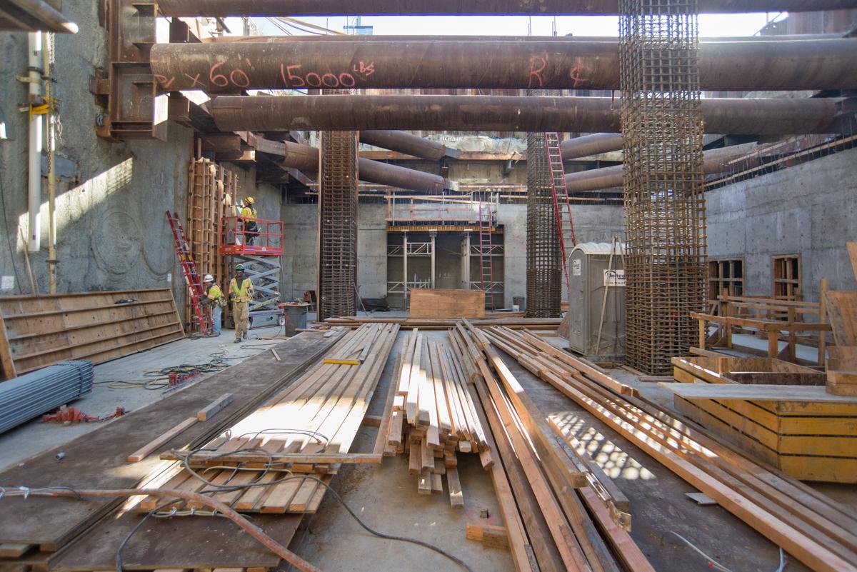 Construction site interior with concrete forms, steel rebar, and structural columns under a steel beam framework.