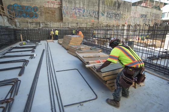 Workers in safety vests install rebar on concrete slab at construction site with metal fencing and urban buildings.