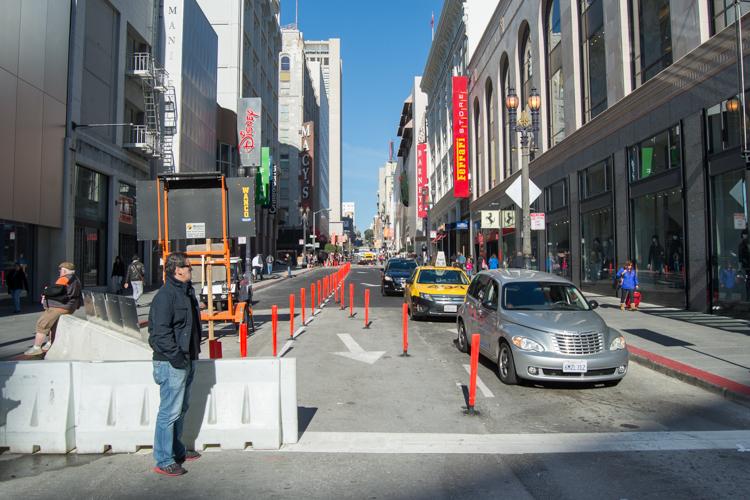 Construction barriers and bollards on Stockton Street in downtown San Francisco with yellow taxis and pedestrians visible.