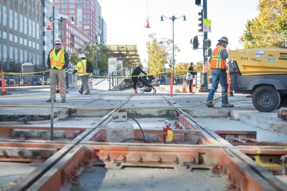 Workers in safety vests install streetcar tracks on an urban street with buildings and a service vehicle nearby.