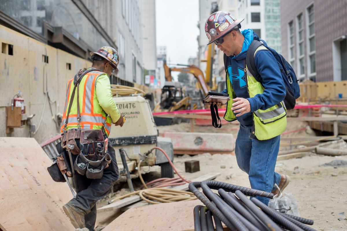 Two construction workers in safety gear discuss plans at a downtown construction site with heavy equipment and buildings visible.