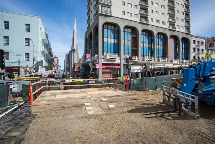 Construction site at Chinatown Station with graded dirt area, equipment, and surrounding San Francisco buildings.