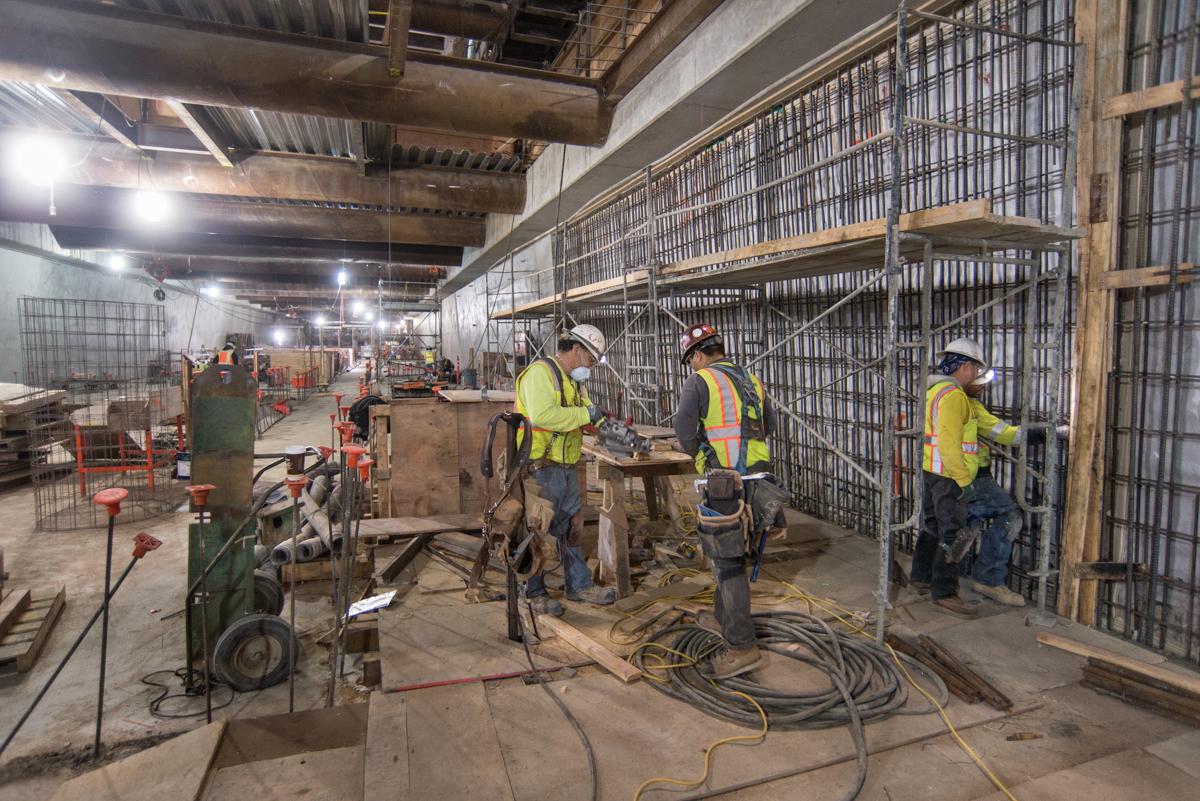 Construction workers in safety vests assemble concrete forms inside a transit station under construction.