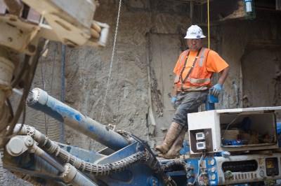 Worker in safety gear operating dewatering well drill equipment on urban construction site.