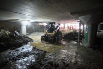 Underground construction site with skid steer loader removing excavated material beneath concrete ceiling.
