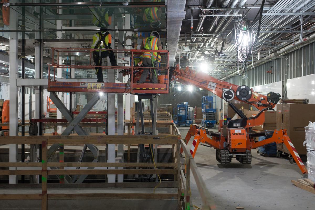 Workers in safety gear operate a cherry picker lift inside an industrial station construction site with exposed framework and equipment.