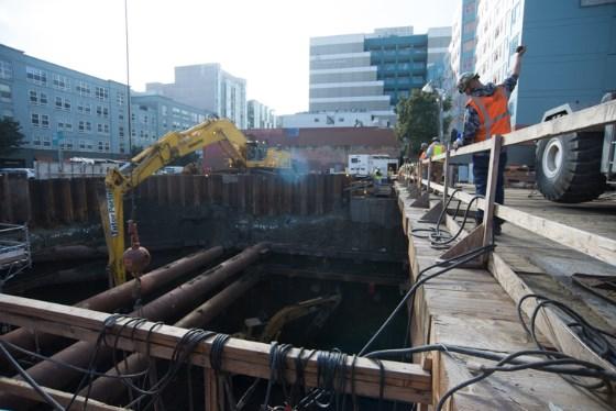 Construction work at underground transit station with yellow excavator lowering equipment into headhouse pit.