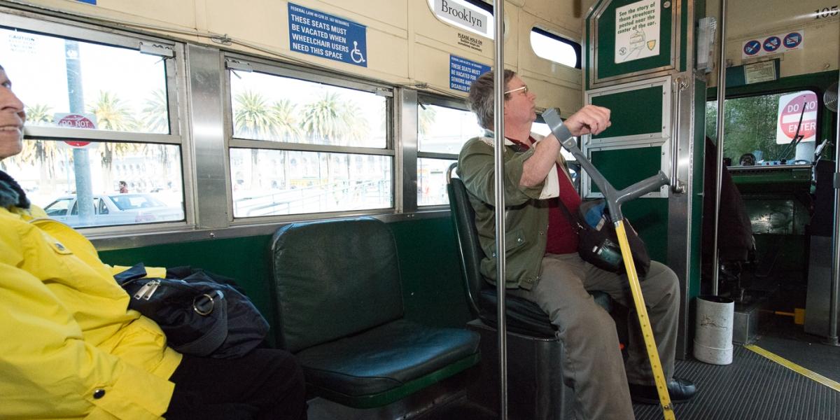Passenger with cane sitting in priority seating area on Brooklyn streetcar near windows.