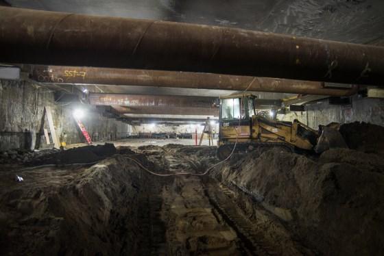 Underground construction site with steel reinforcement beams, excavated soil, and heavy equipment beneath concrete structure.