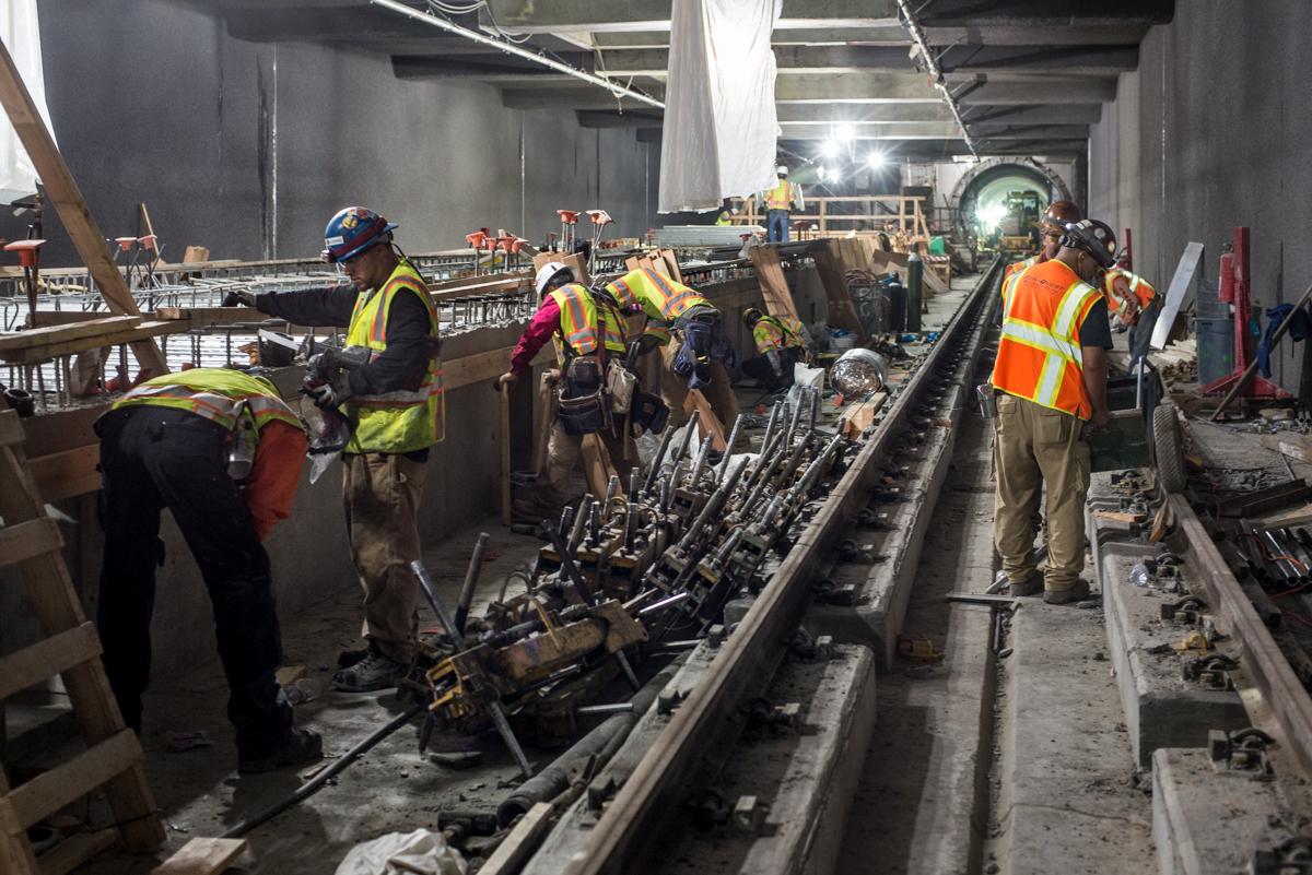 Construction workers in safety vests install rail infrastructure and formwork under a concrete overpass at a transit station.