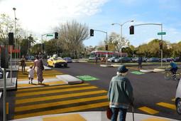 Pedestrians crossing at yellow striped crosswalk with traffic signals and vehicles at intersection.