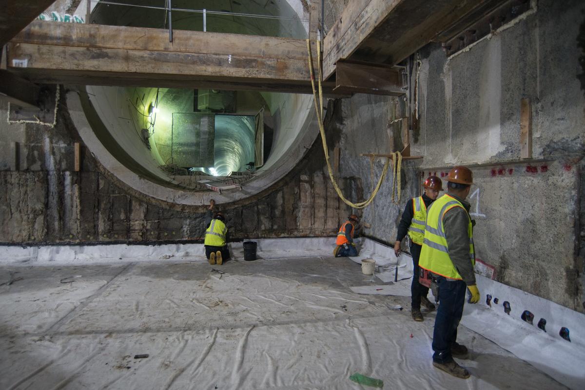 Workers in safety vests inside underground transit station construction area with large circular tunnel opening above.