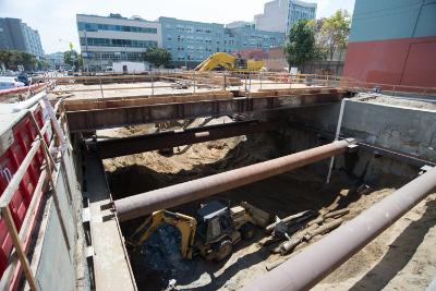 Construction excavation pit with temporary steel supports and large pipes, urban setting with buildings in background.