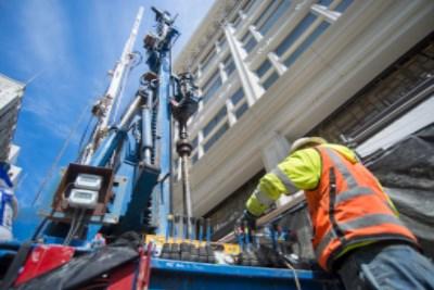 Worker in safety vest operating jet grouting drill rig on urban street near building.