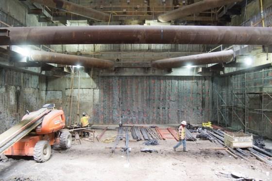 Industrial construction site with orange machinery, metal framework, and rebar installation work underway in underground station headhouse.