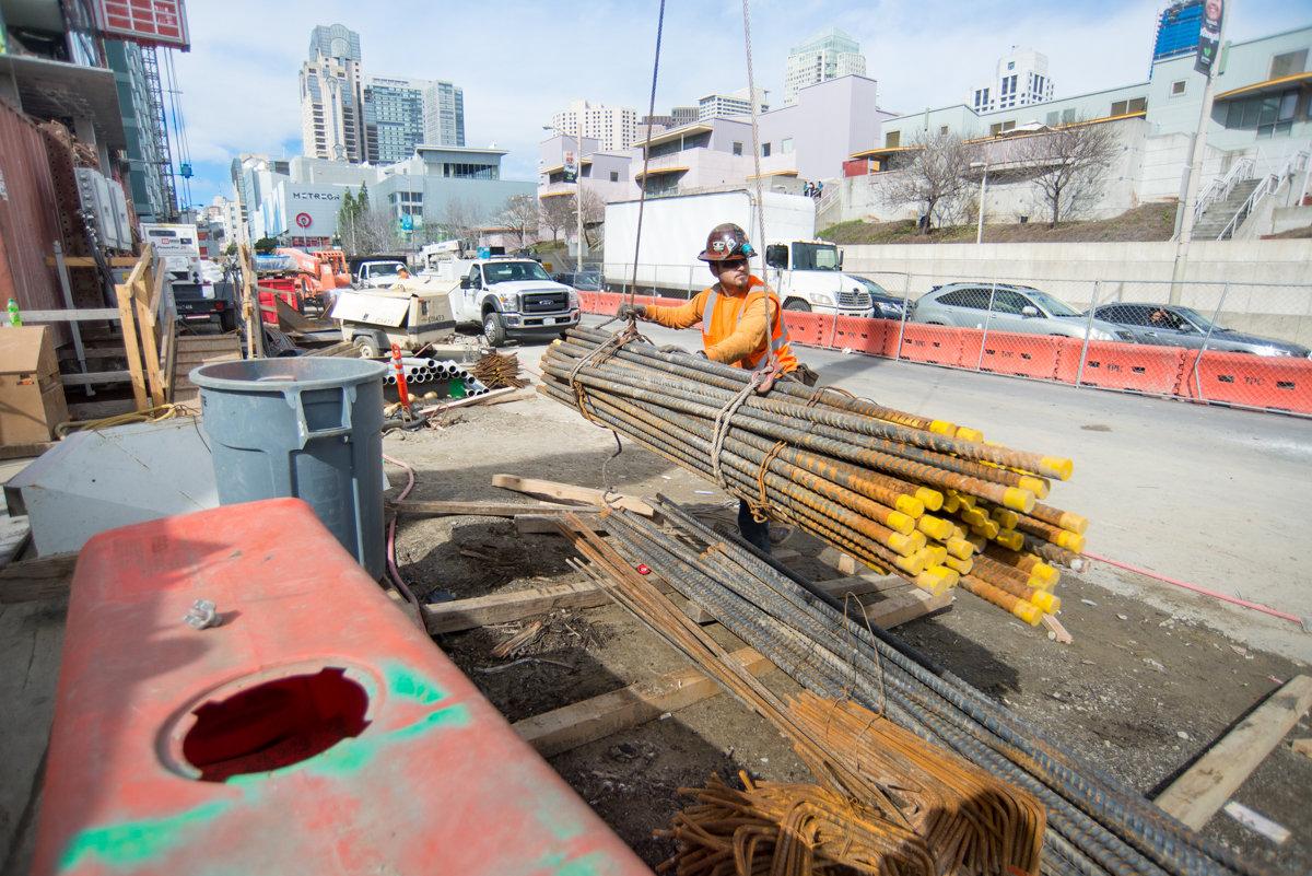 Worker guides rebar bundle during crane lift at transit station construction site.