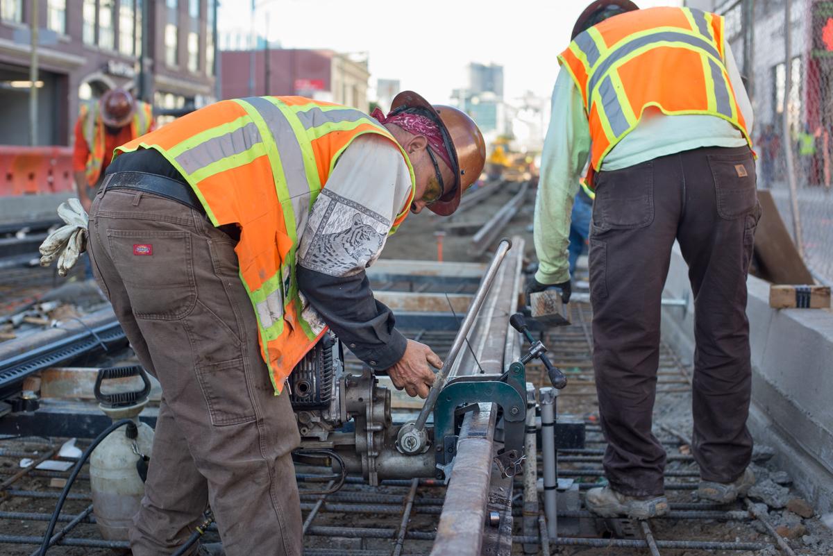 Workers in safety vests drill holes through steel rails on a transit construction site.