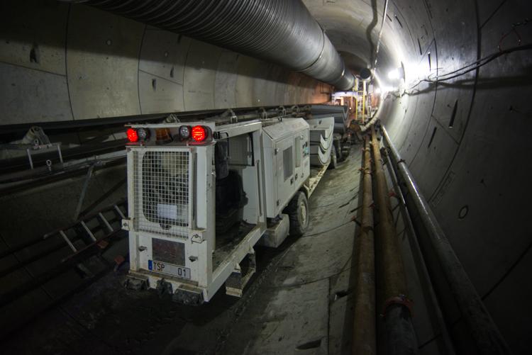 Construction vehicles with red lights inside concrete tunnel with metal pipes overhead.