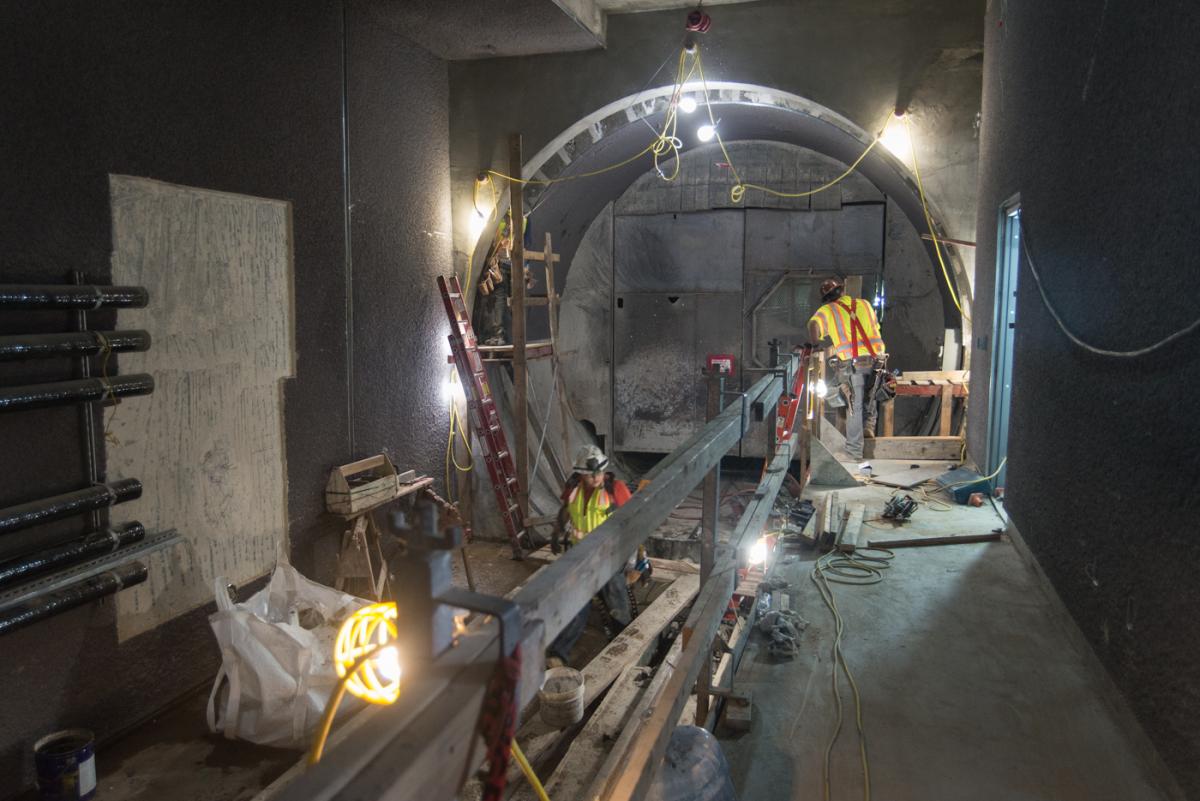 Construction workers in safety gear work on tunnel infrastructure with lighting and equipment visible in an underground transit station.