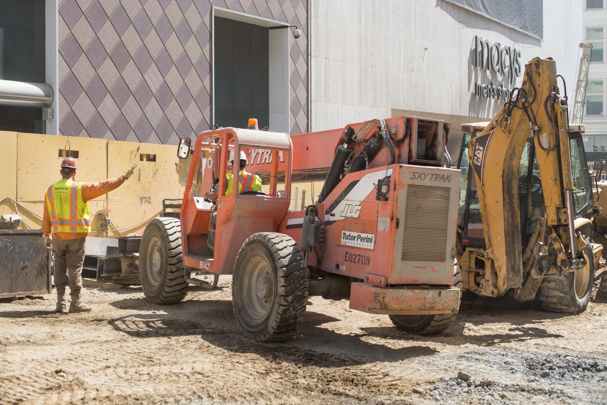 Construction workers coordinate heavy equipment and excavated materials at an active transit station construction site.