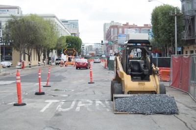Construction equipment compacting gravel on urban street with traffic cones and utility work barriers.