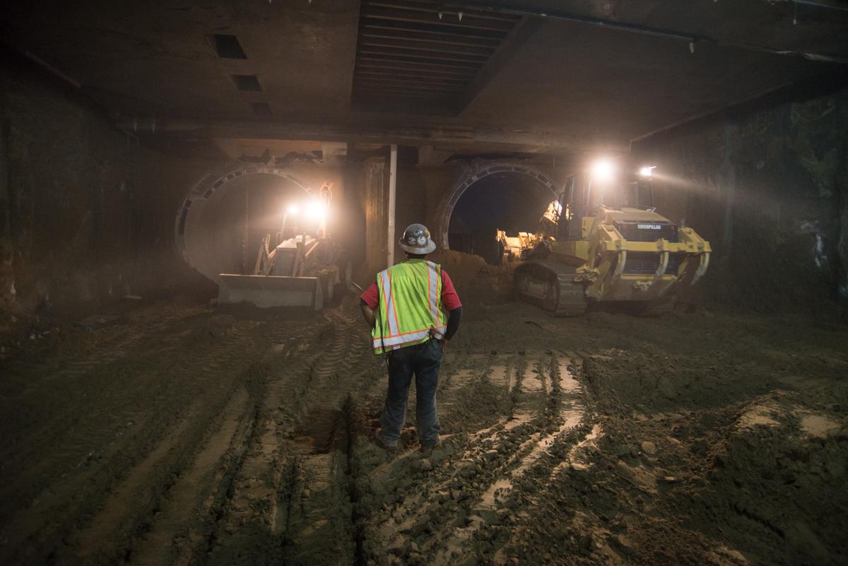 Worker in safety vest stands in underground tunnel with construction equipment and lighting.