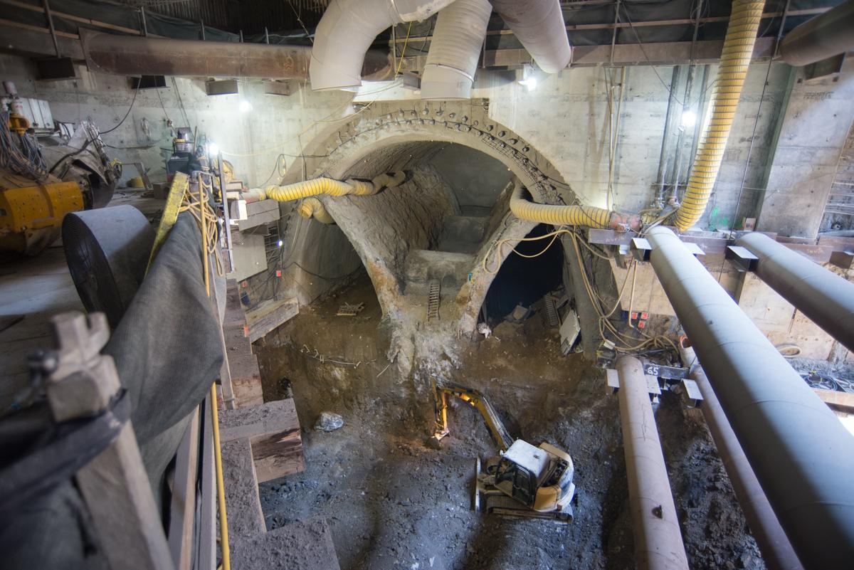 Large concrete tunnel entrance under construction with yellow hoses and equipment inside excavated underground chamber.
