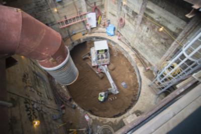 Aerial view of underground transit construction site with excavation pit, equipment, and surrounding buildings.
