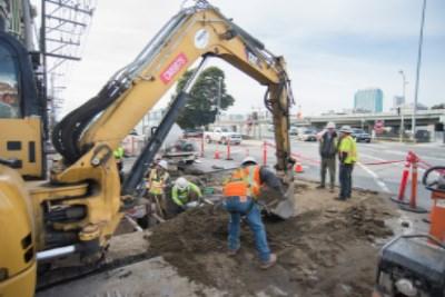 Yellow excavator operates in street trench with construction workers and equipment nearby.
