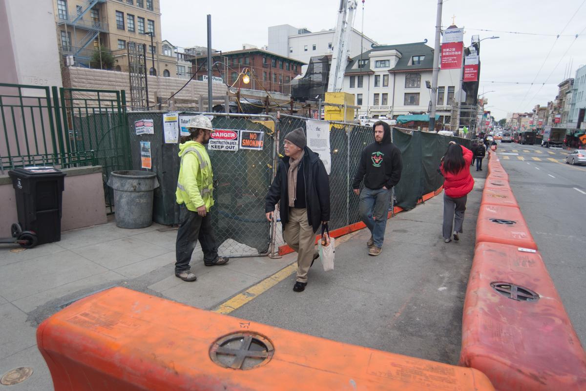 Pedestrians walk along temporary orange barriers on a city street near construction fencing and equipment.