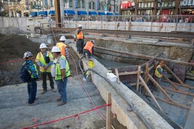 Construction workers in safety vests work on underground infrastructure at Ellis and Market Street.