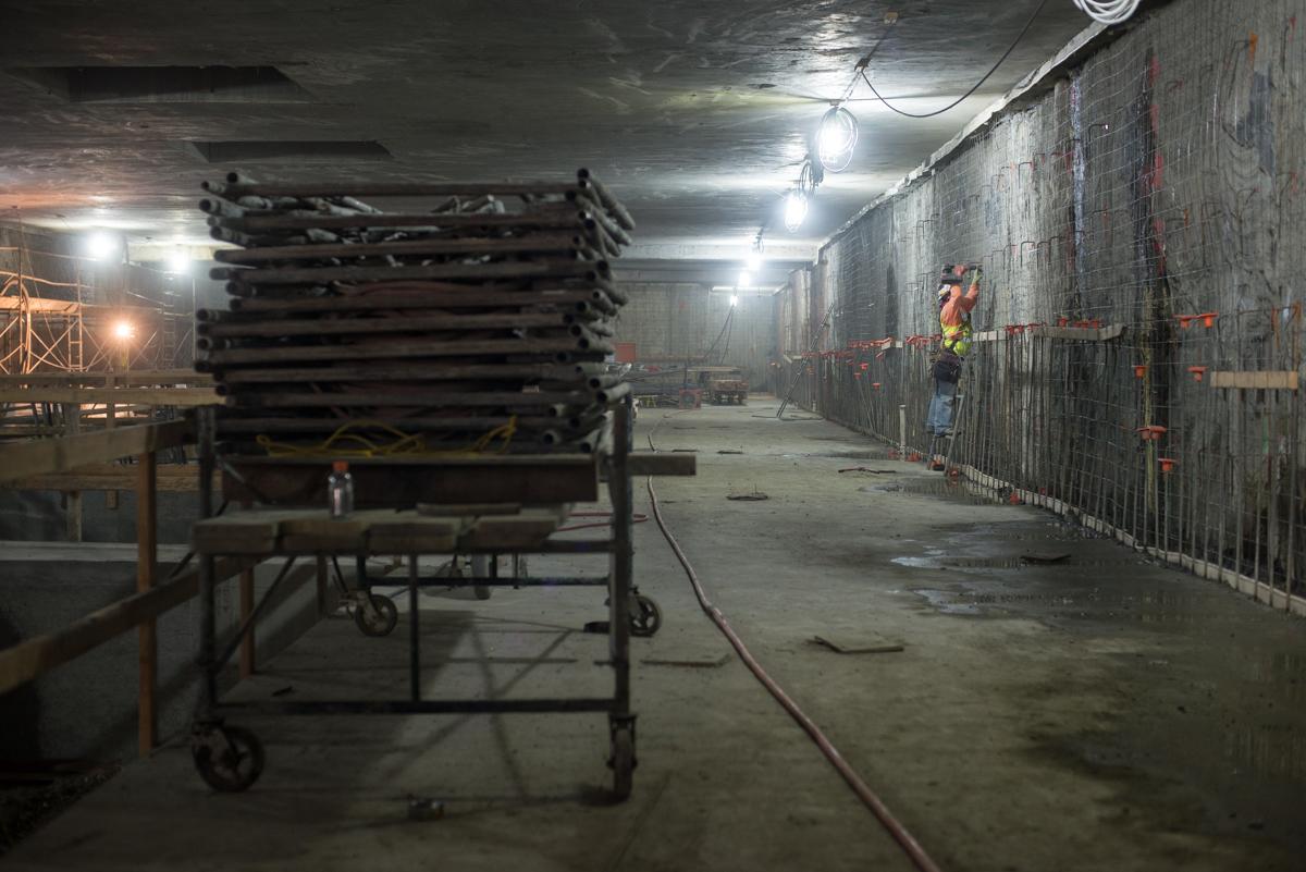 Underground construction site with stacked metal pallets, support structures, temporary fencing, and workers in safety gear installing utilities and rebar.