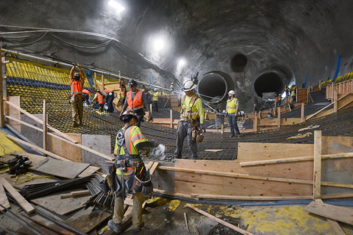 Workers in safety gear construct formwork and rebar inside underground Chinatown Station tunnel during transit construction.
