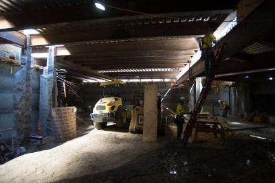 Excavator with slotted bucket sifting material inside industrial construction site with overhead lighting and concrete pillars.