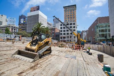 Construction site with yellow excavator and heavy equipment on sandy ground, downtown buildings in background