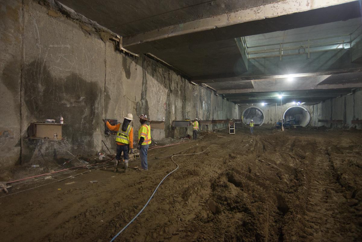Workers in safety vests conduct construction work inside a large underground transit tunnel with exposed concrete walls and dirt flooring.