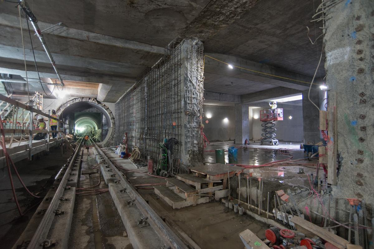 Underground transit tunnel under construction with exposed concrete, steel reinforcement, and work equipment visible.