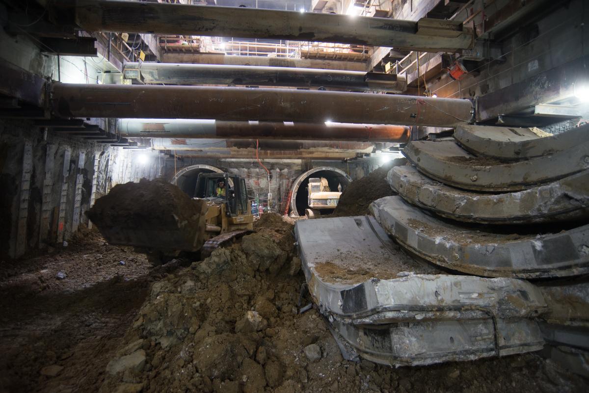 Underground construction site with stacked concrete tunnel segments and excavation equipment beneath steel beams.