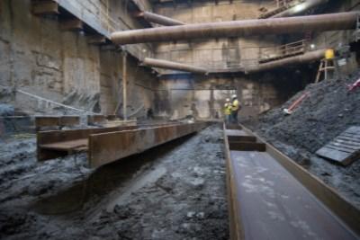 Underground construction site showing steel I-beams on slurry walls, railroad tracks, and excavated earth beneath Stockton Street.